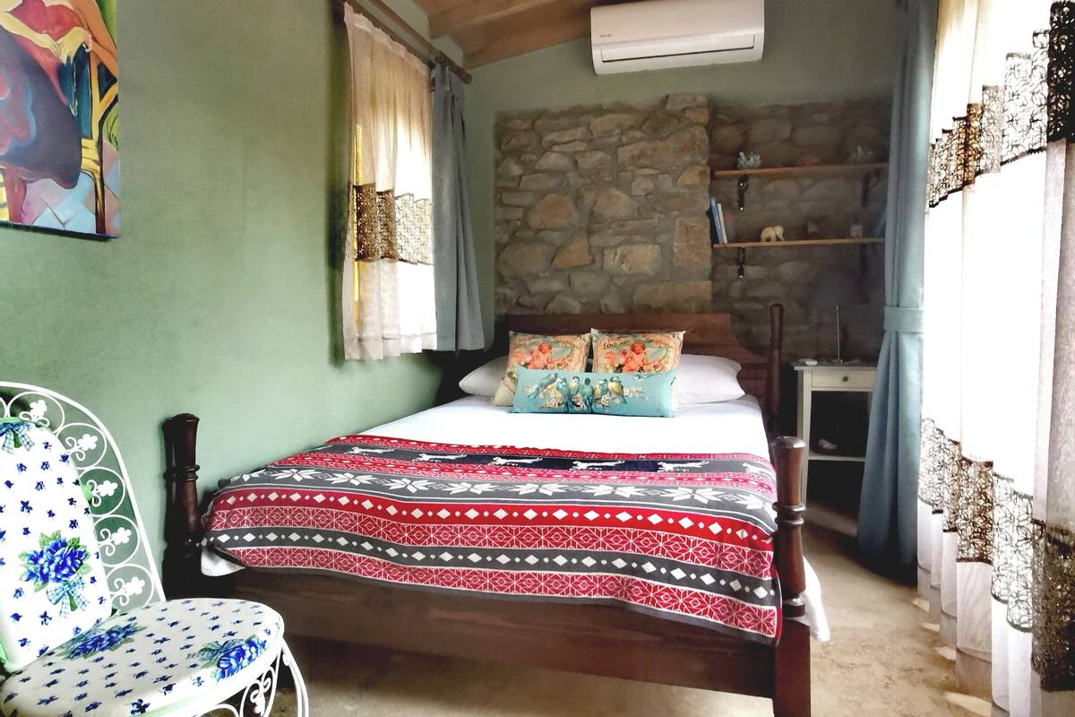 A bedroom with a wooden ceiling, a bed with patterned bedding, a chair with a floral design, a stone wall, and an air conditioning unit.
