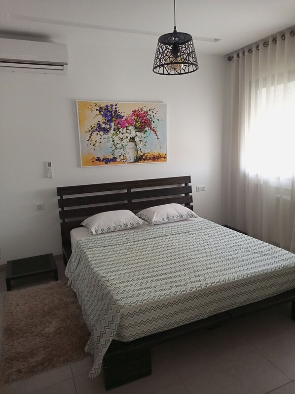 Une chambre à coucher avec un lit, une table de chevet, un radiateur, un tableau et une fenêtre avec des rideaux.