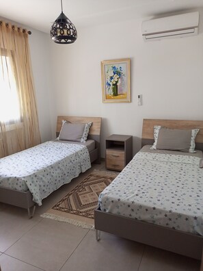 Une chambre avec deux lits simples, une table de chevet, une petite armoire, un luminaire suspendu, un climatiseur fixé au mur et un tableau encadré.