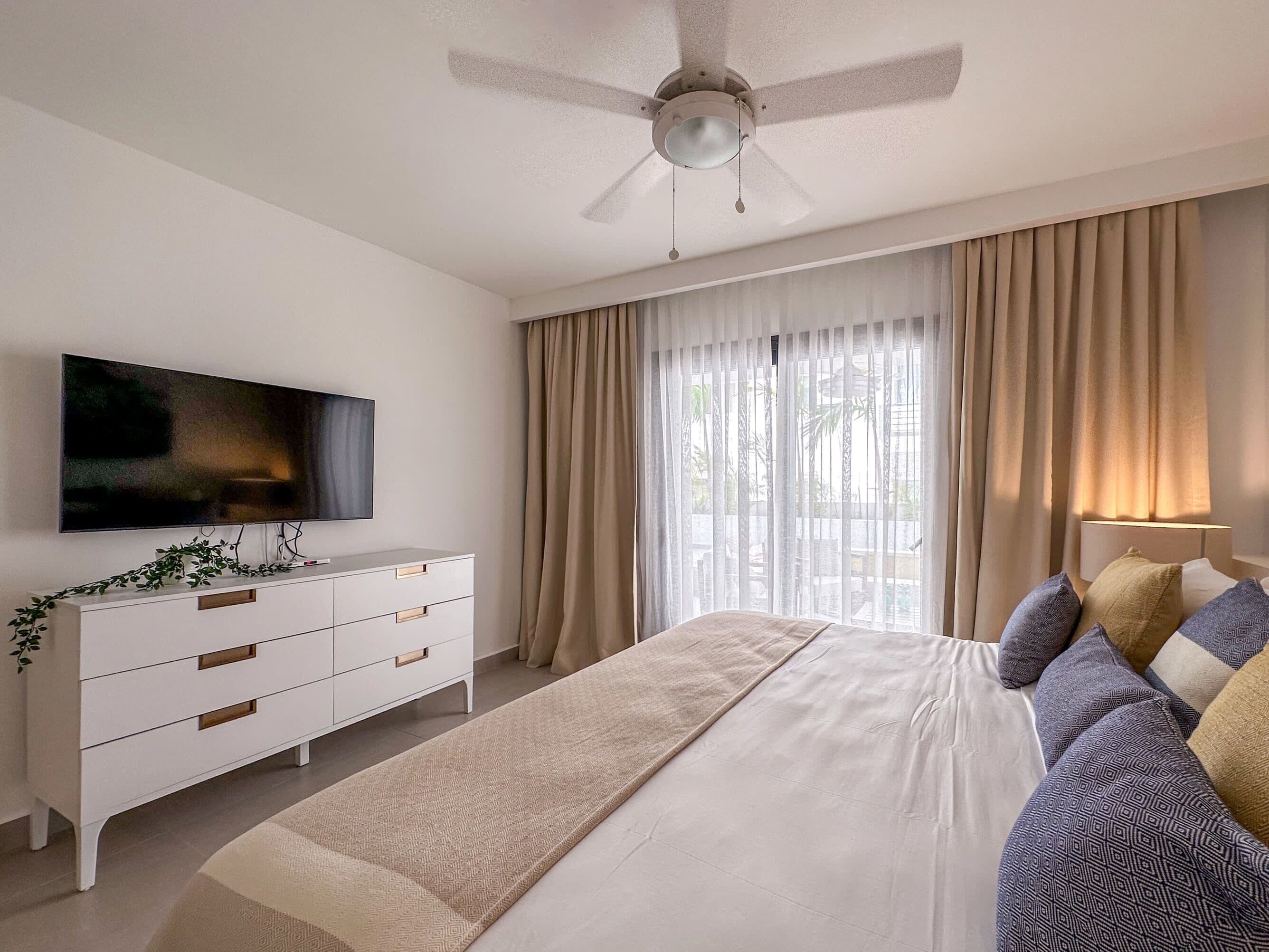 A bedroom with a bed, a TV, a ceiling fan, and a window with curtains.