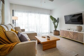 Una sala de estar moderna con sofá, mesa de centro, televisor y un gran ventanal con cortinas translúcidas.