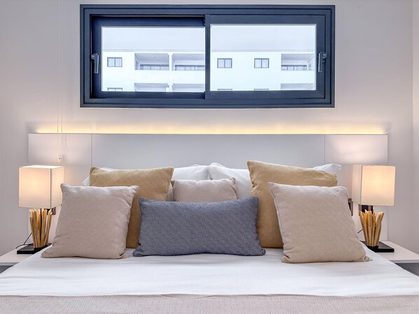 A neatly arranged bed with multiple pillows, two bedside lamps, and a window above.