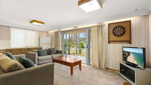 A spacious living room with a sofa set, a coffee table, a flat-screen TV, a dartboard, and a view of the outdoors through large glass doors. - Minerva Beach House by Experience Jervis Bay (Vincentia)