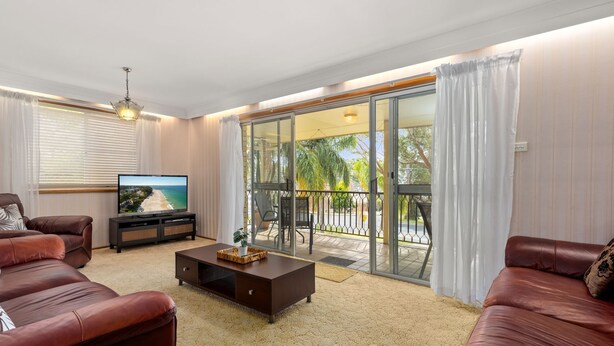 A living room with a TV, leather sofas, a coffee table, and a balcony with a view of the beach. - Minerva Beach House by Experience Jervis Bay (Vincentia)