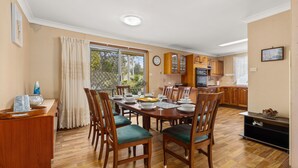 A dining area with a wooden table set for four, surrounded by chairs with green cushions. A kitchen with wooden cabinets and a refrigerator is visible in the background. - Minerva Beach House by Experience Jervis Bay (Vincentia)