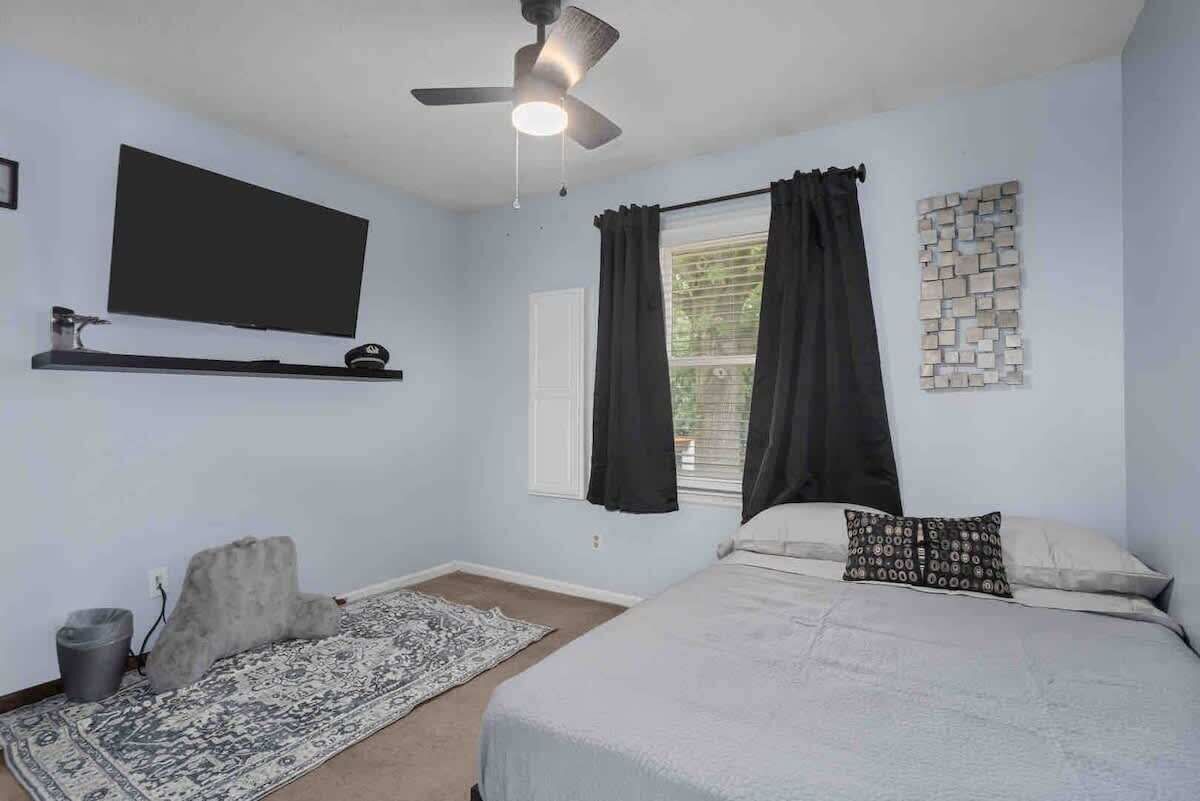 Un dormitorio con cama, ventilador de techo, televisor montado en la pared, ventana con cortinas, alfombra y un pequeño estante con un objeto decorativo.