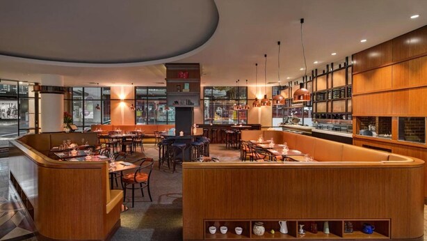 Un restaurante moderno con barra central, diversas opciones de asientos y vistas a la calle a través de amplios ventanales.
