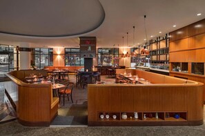 A modern restaurant with a central bar area, multiple seating options, and a view of the street through large windows.