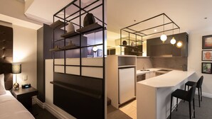 Una habitación de hotel moderna con una kitchenette separada, una cama con mesita de noche y un escritorio con lámpara.