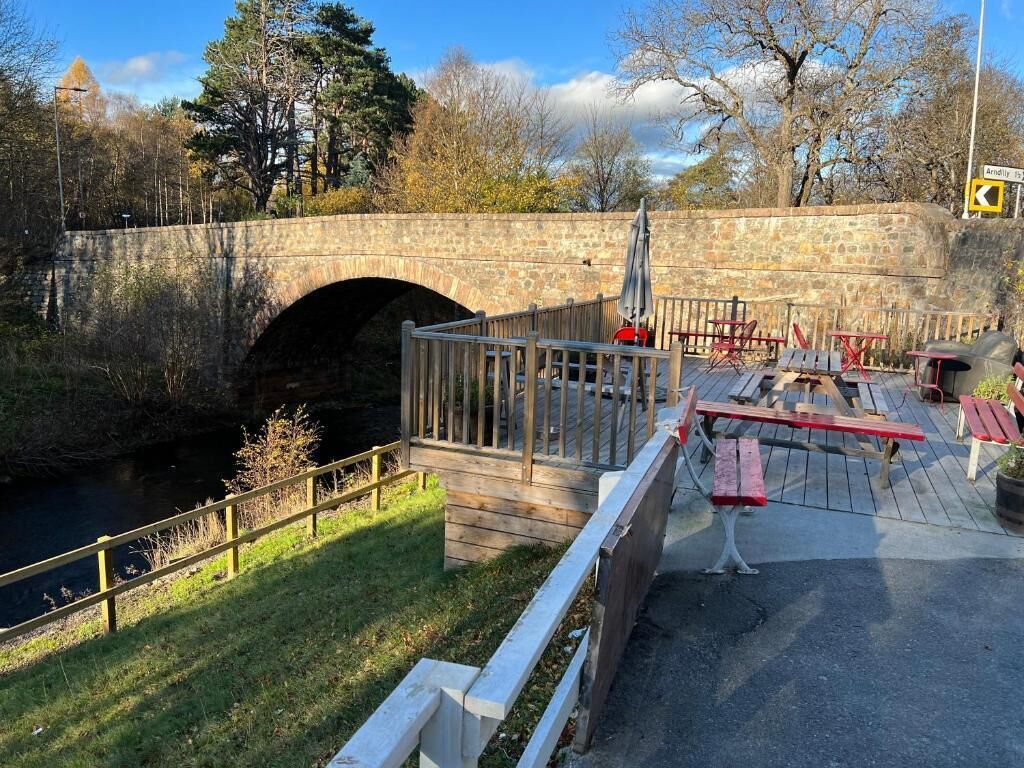 Eine Steinsbrücke über einem Fluss, ein Holzdeck mit Bänken und Tischen sowie eine Rasenfläche mit Bäumen.