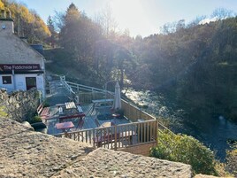 El hostal Fiddichside con un porche con vistas a un río, paredes de piedra y vistas del bosque circundante.