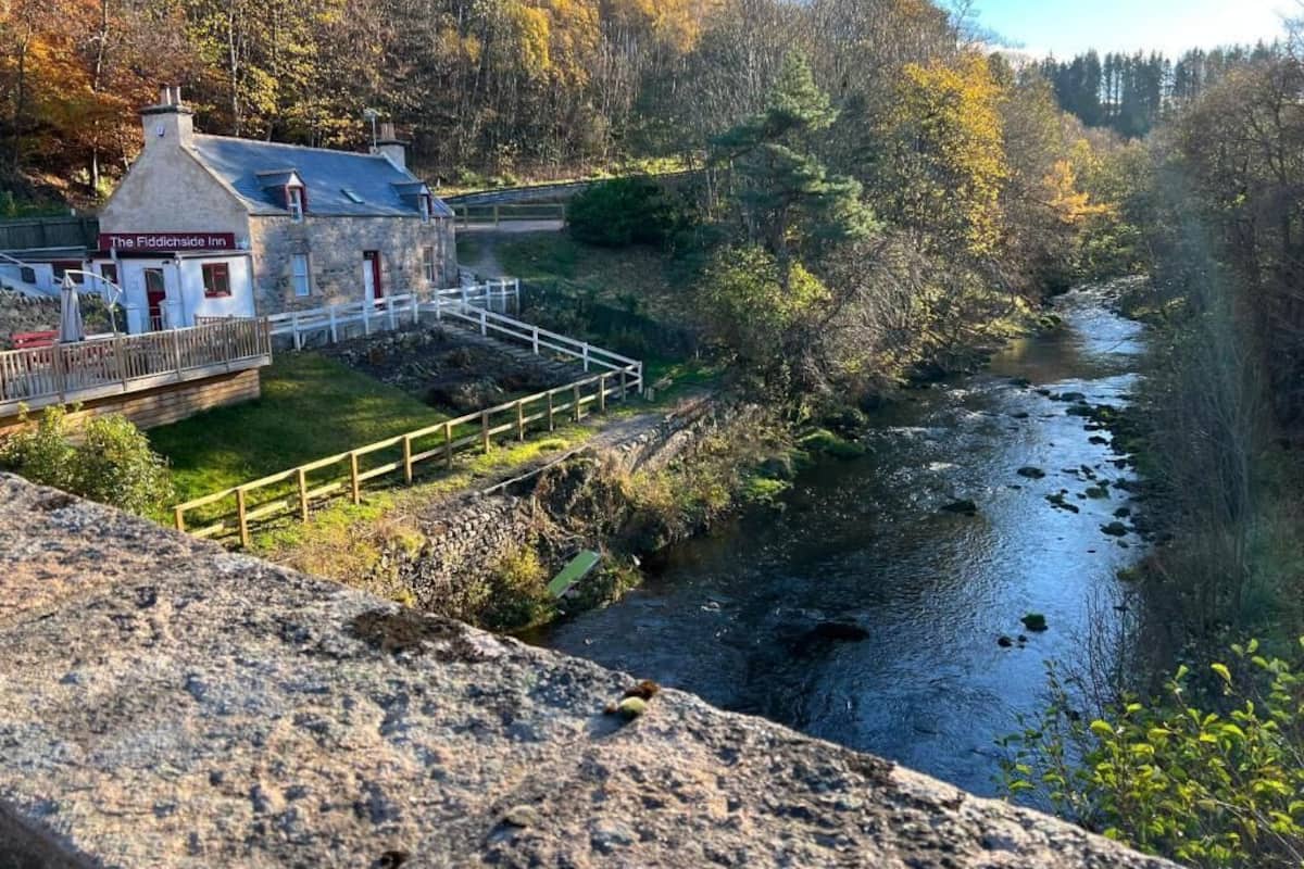 The Fiddichside Inn, a stone building with a white facade and a red door, located beside a river with a wooden bridge leading to it.