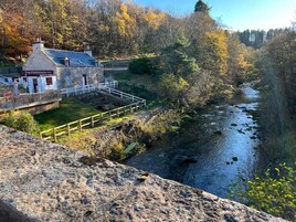 El Inn Fiddichside, un edificio de piedra con fachada blanca y puerta roja, situado junto a un río con un puente de madera que lo atraviesa.