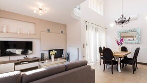 A modern living room with a sofa, TV, fireplace, dining table with chairs, and a chandelier. - Peris 2 Luxury Villa In Chania, Private Pool (Daratsos)