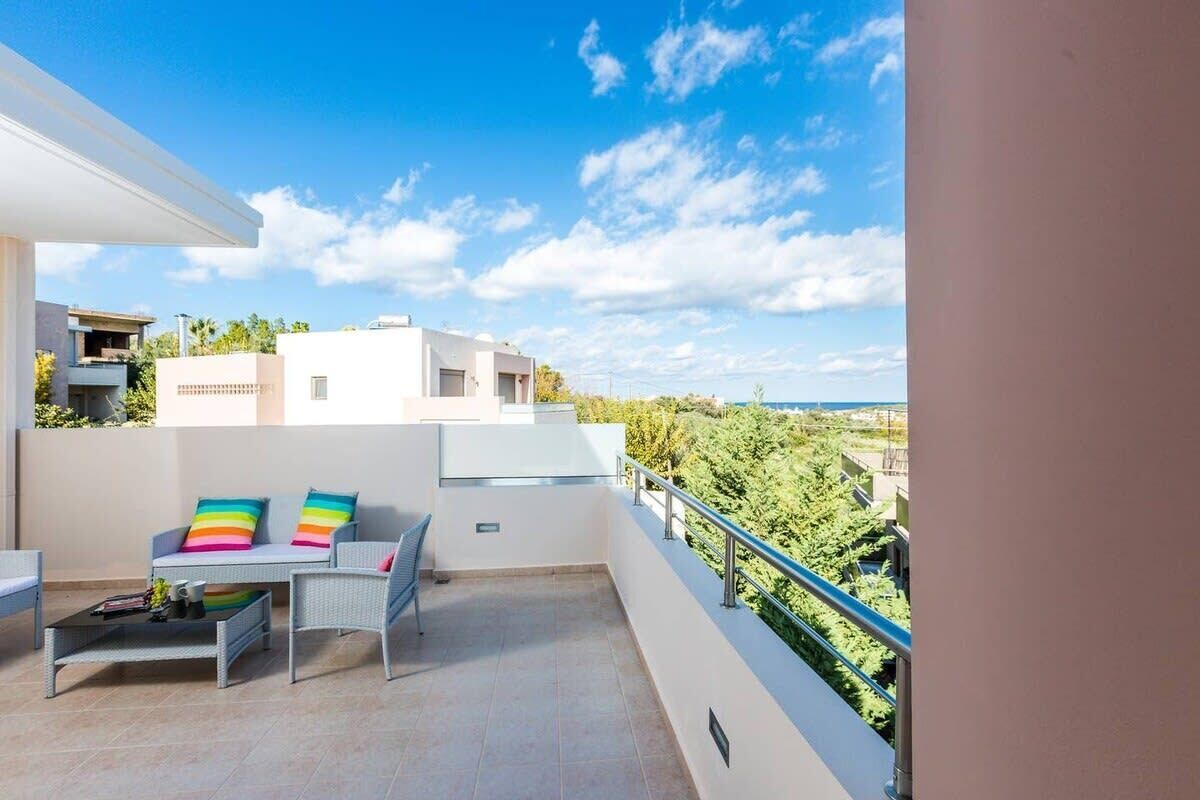 A balcony with a white railing, a table with colorful cushions, and a view of buildings and greenery.