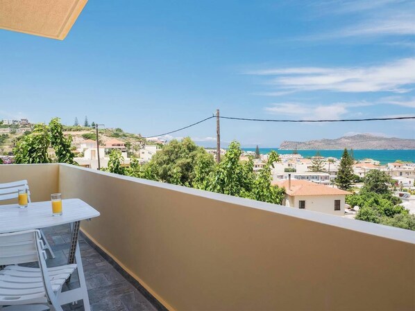 A balcony with a table and chairs, overlooking a coastal town with buildings and a clear sky. - Romantic Corner - Anoula Osase Sea View Maisonette (Stalos)