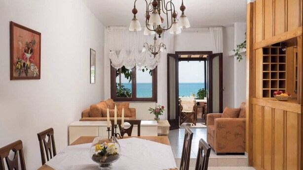 Un comedor con una mesa de madera, sillas y una araña. Hay un sofá y vistas al mar a través de una ventana.