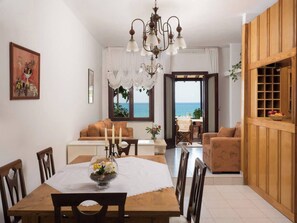 A dining area with a wooden table, chairs, and a chandelier. There is a sofa and a view of the sea through a window. - Romantic Corner - Anoula Osase Sea View Maisonette (Stalos)