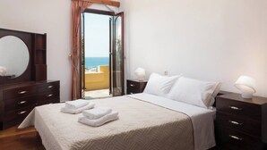 Un dormitorio con cama, mesitas de noche, un espejo y vista al mar a través de una puerta abierta.