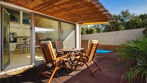 En udendørs terrasse med et træbord og stole, et stråtækt tag og en glasdør, der fører ind til et køkken.