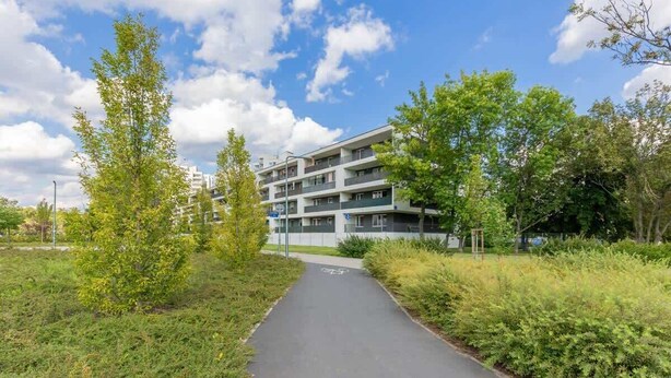 A pathway leading to a multi-story residential building with balconies, surrounded by greenery and trees.