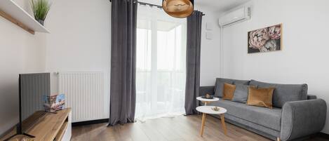 A modern living room with a grey sofa, a small round table, a wooden floor, and a large window with grey curtains.