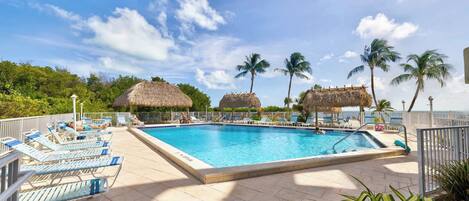A swimming pool with lounge chairs and thatched-roof structures, surrounded by palm trees and a clear sky.