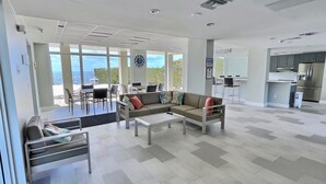 A spacious living area with a sofa, coffee table, and dining area. There is a kitchen with modern appliances and a view of the ocean through large windows.