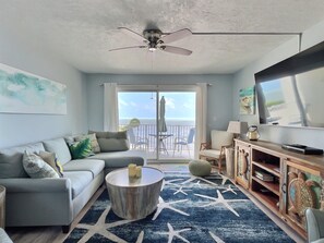 A living room with a sofa, coffee table, TV, and a balcony with a view of the ocean.