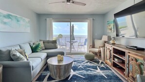 A living room with a sofa, coffee table, TV, and a balcony with a view of the ocean.