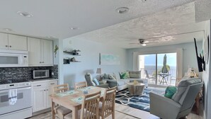 A modern kitchen with white cabinets, a microwave, and a stove. There is a dining area with a wooden table and chairs. A gray sofa and armchair are in the living area, which has a large window with a view of the ocean.