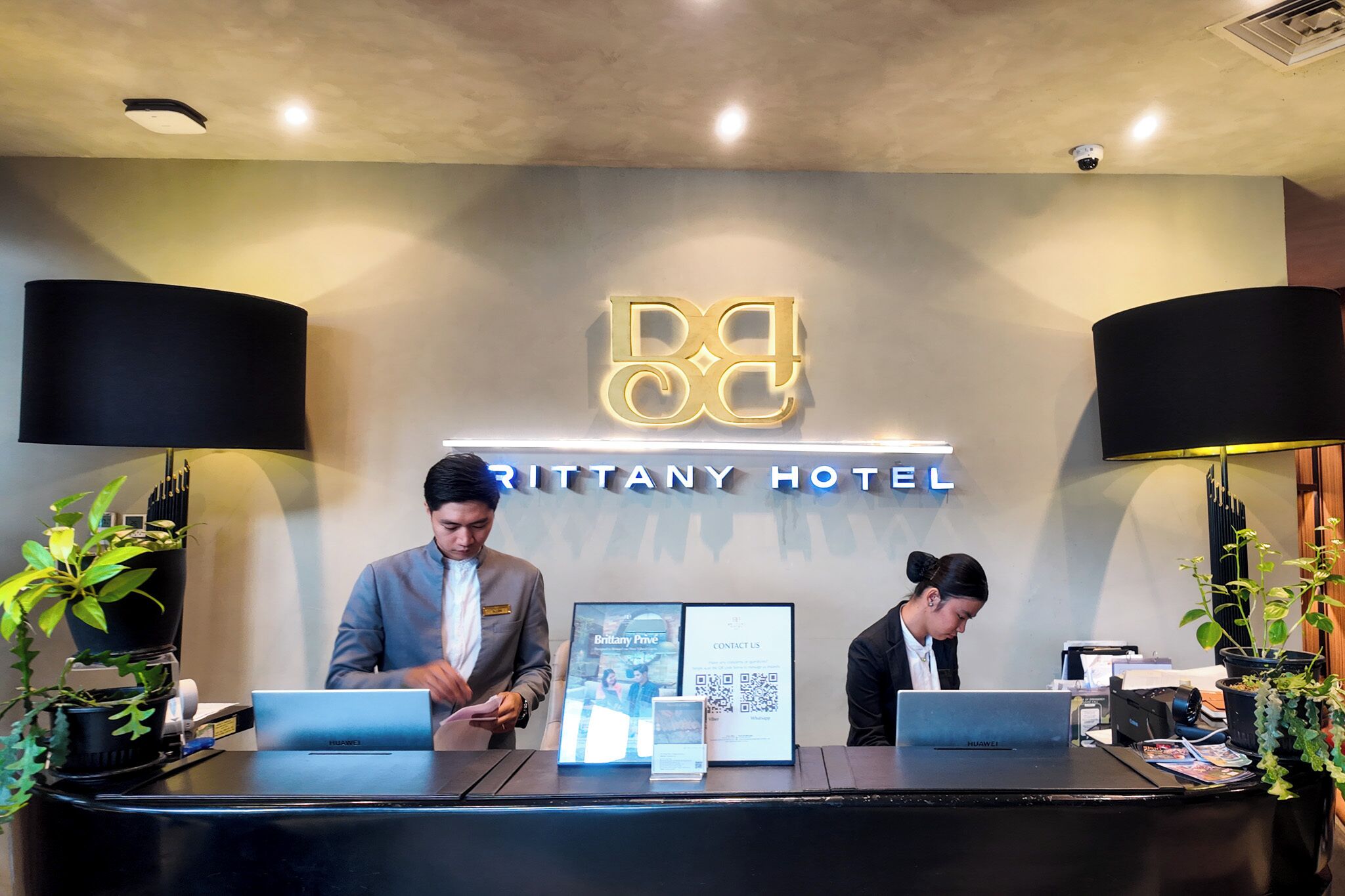 A hotel reception desk with a sign reading 'Brittany Hotel,' two staff members, a laptop, a framed photo, and a QR code.