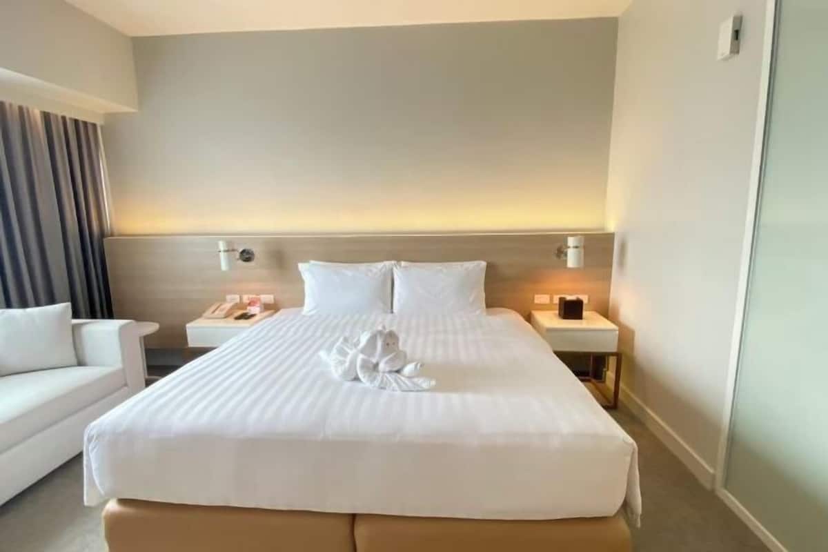 Una habitación de hotel con una cama grande, una mesita de noche, un sillón blanco y una lámpara de pared.