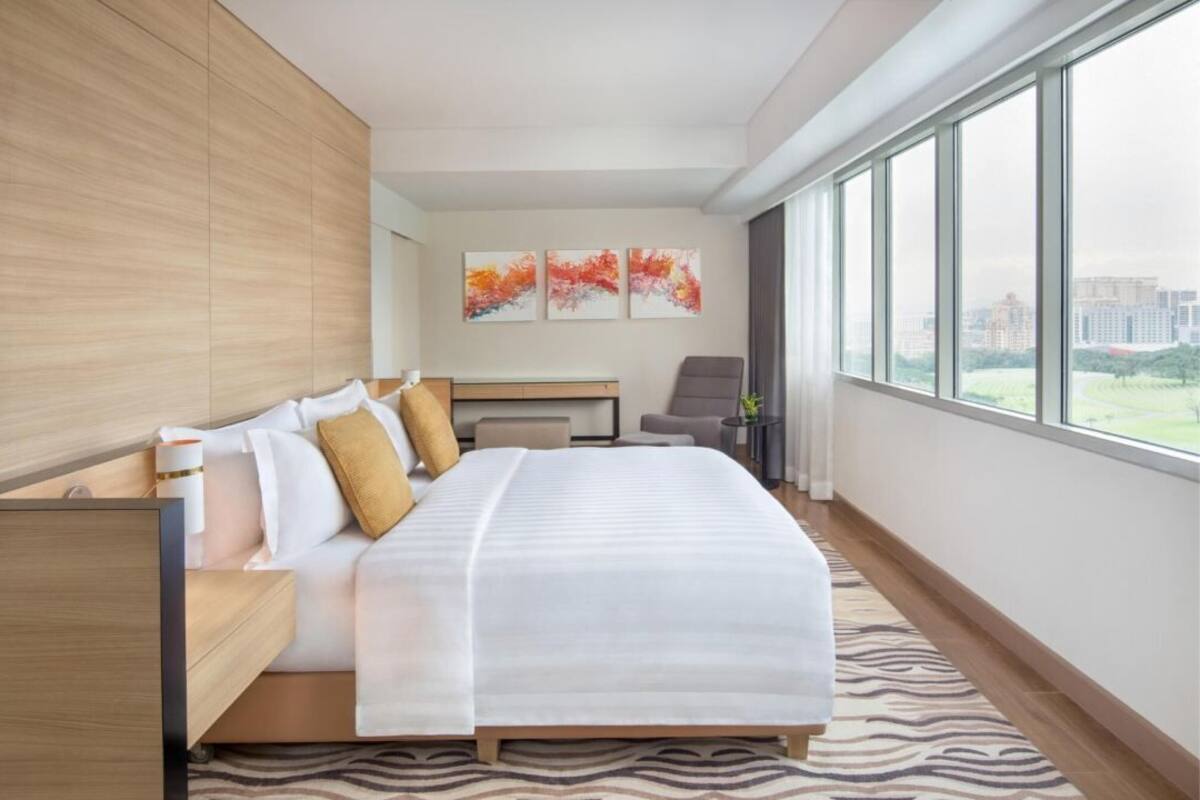 Habitación de hotel moderna con una cama grande, un escritorio, una silla y vistas a la ciudad a través de amplios ventanales.