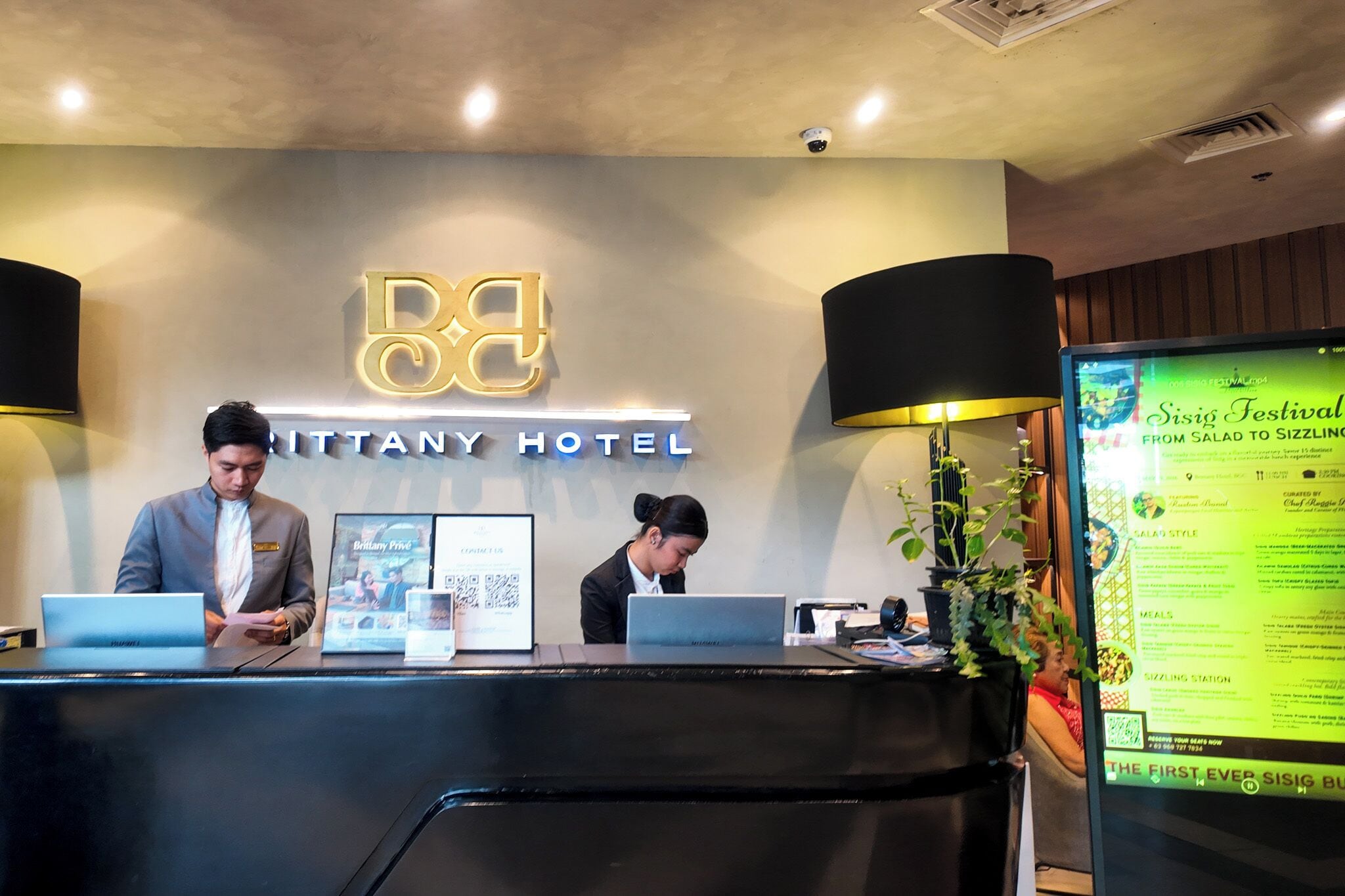 A reception desk with two staff members, a digital menu board, and a sign for the Bittany Hotel.