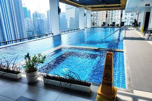An indoor swimming pool with city views, potted plants, and a caution wet floor sign. - Brittany Hotel BGC (Taguig)