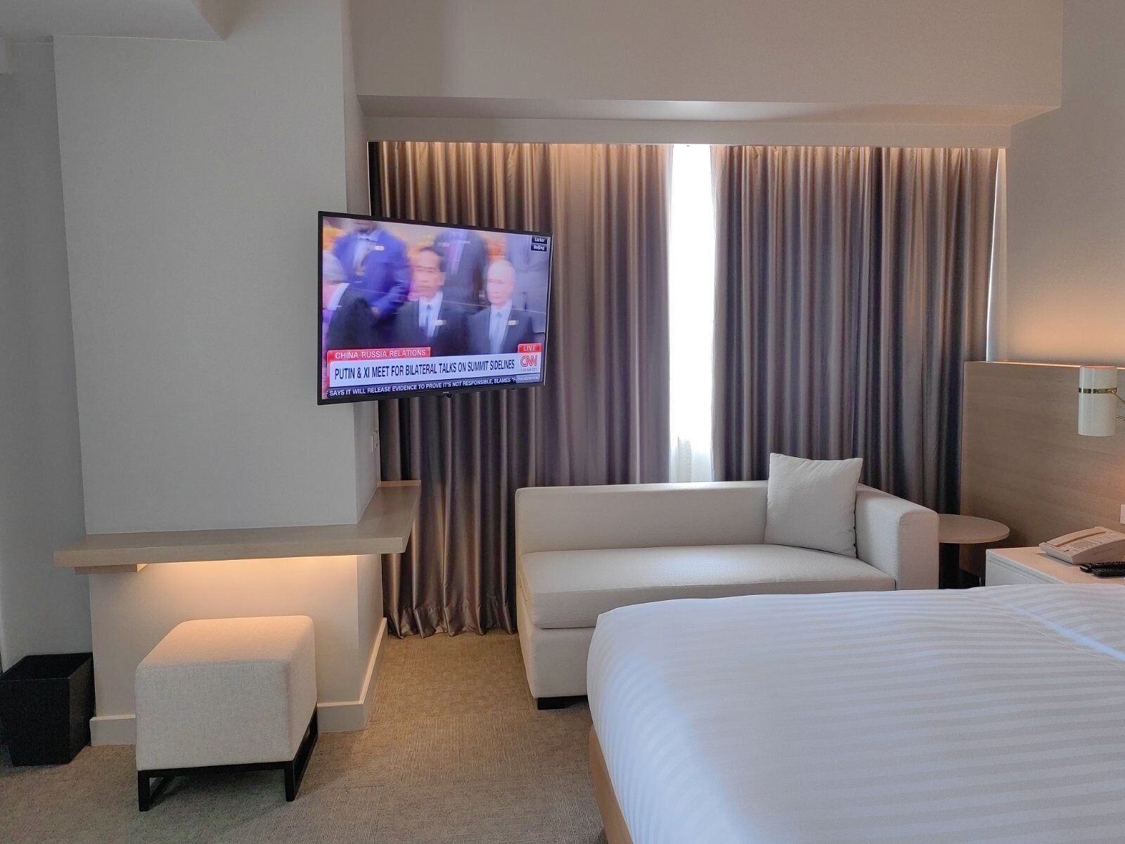 Habitación de hotel con una cama grande, un sofá, un televisor montado en la pared y una ventana con cortinas.