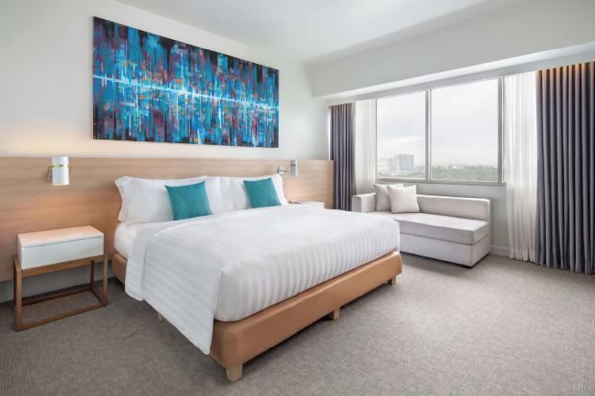 A hotel room with a large bed, a bedside table, a sofa, a window with curtains, and a city view.