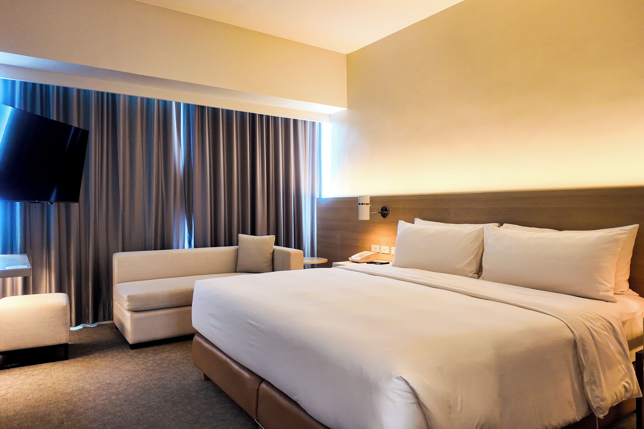 A hotel room with a large bed, a flat-screen TV mounted on the wall, a seating area with a sofa and armchair, and a window with curtains.