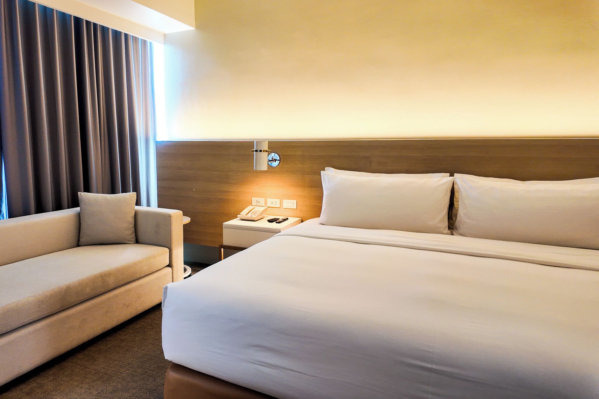 Une chambre d’hôtel avec un lit, un canapé, une table de chevet avec un téléphone et une lampe fixée au mur.