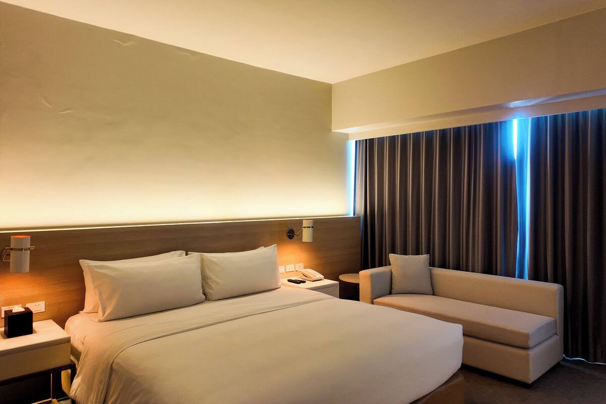Habitación de hotel con cama, mesita de noche, lámpara, teléfono y sofá.