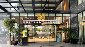 El exterior del Hotel Brittany, con una fachada moderna de vidrio, grandes macetas con plantas y un área de estar al aire libre visible.