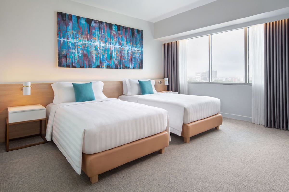Habitación de hotel con dos camas, un cuadro abstracto grande en la pared, vista a la ciudad a través de la ventana y una mesita de noche con una lámpara.