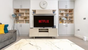 Una sala moderna con un televisor de pantalla plana montado en un mueble blanco, un reloj encima y una estantería con diversos objetos.