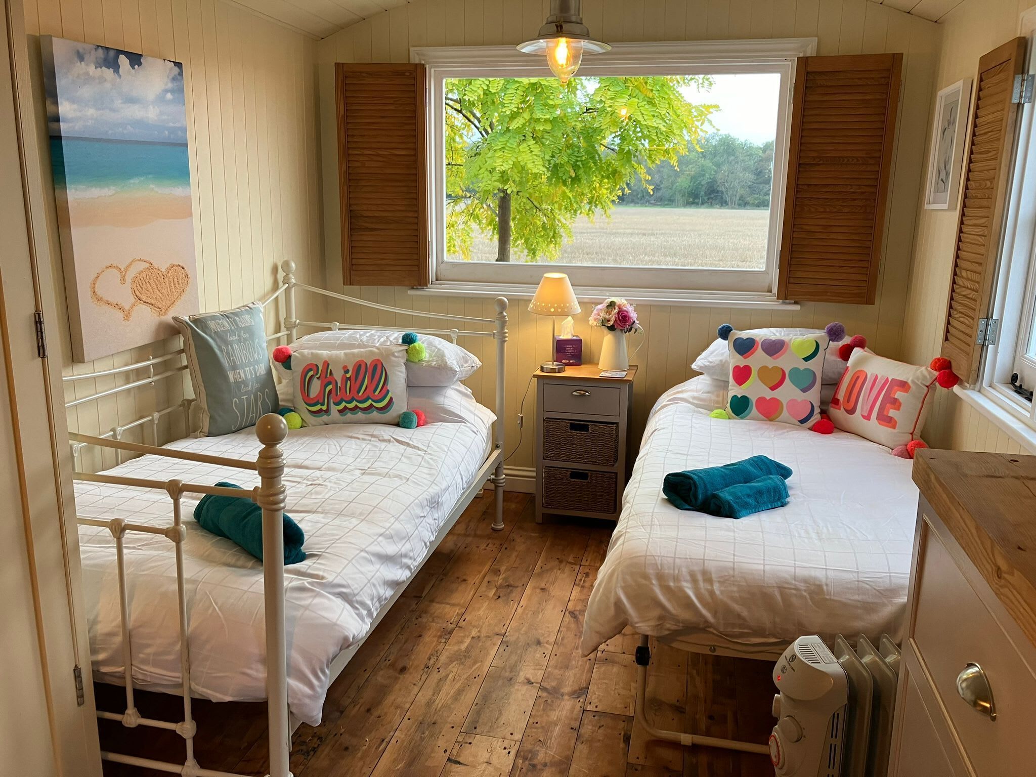 A cozy bedroom with two beds, a nightstand with a lamp, a window with a view of trees, and a painting on the wall.