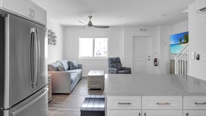 A modern kitchen with a refrigerator, a dining area with a table and chairs, a sofa, and a ceiling fan.