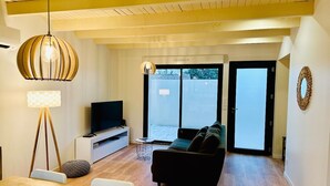 A modern living room with a wooden ceiling, a black sofa, a TV on a white console, and a large window with a view of the outdoors. - Le Grand *** - 3 bedrooms, all new & all inclusive (Montauban)
