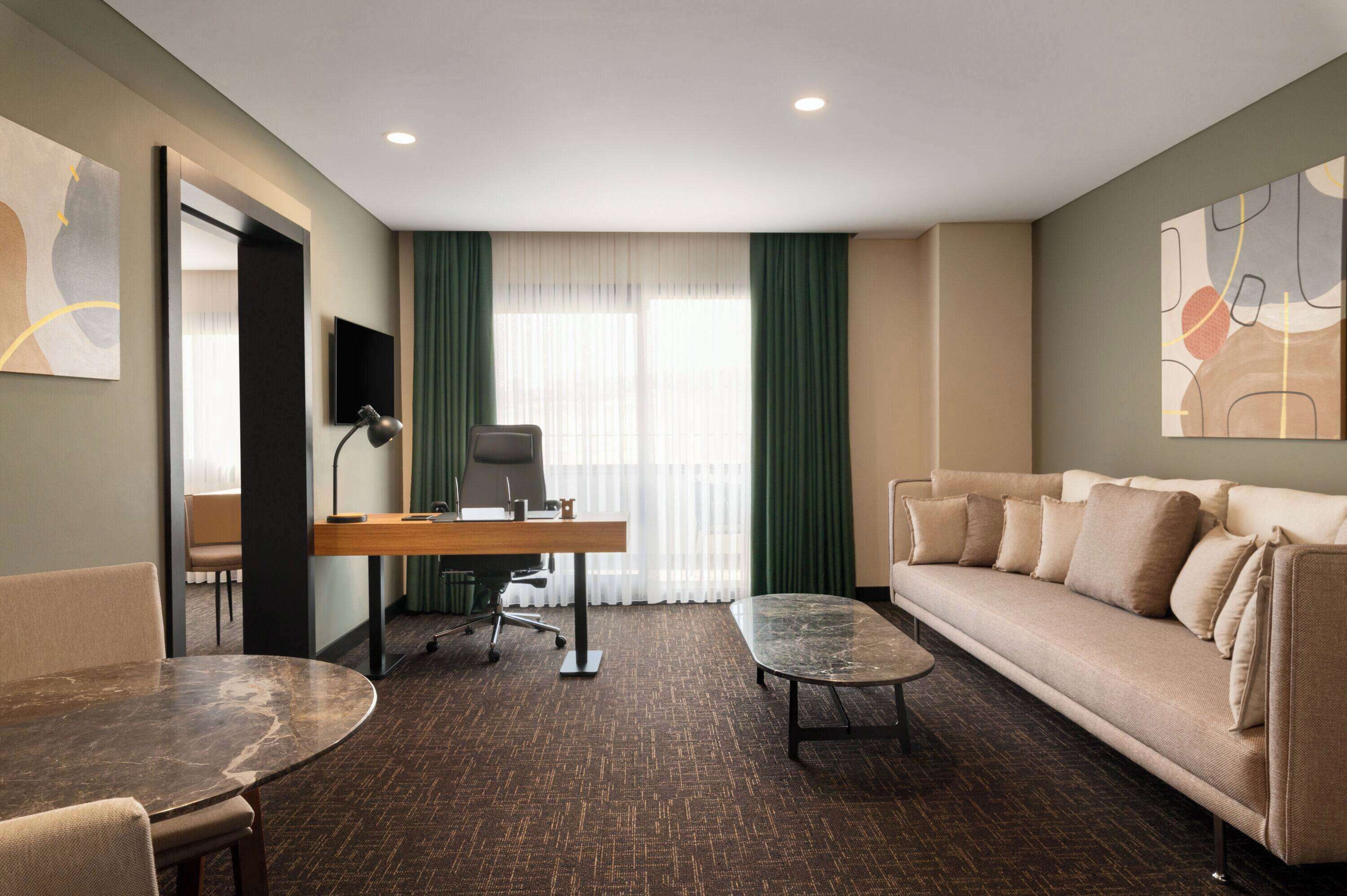 Habitación de hotel con sofá, escritorio, silla, una mesita y televisor en la pared.