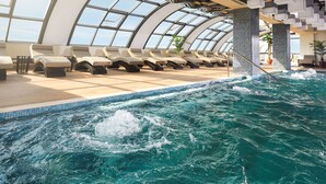 Un espace piscine intérieur moderne, doté de chaises longues et d’un grand plafond en verre transparent.