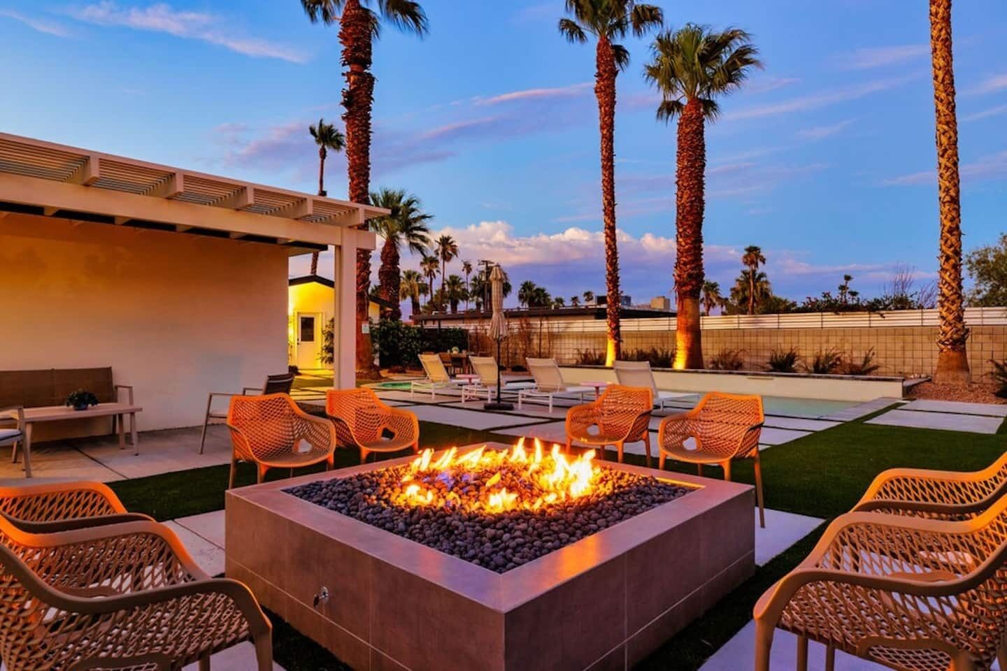 A fire pit surrounded by wicker chairs, a pool, and palm trees.